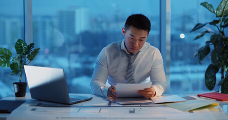 Asian young businessman working in corporate business office department examining financial papers doing paperwork alone overworking.