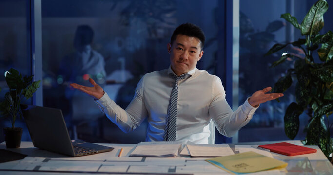 Portrait Of Chinese Adult Business Manager Doing Financial Paperwork At Corporate Desk Shrugging Shoulders In Hesitation.