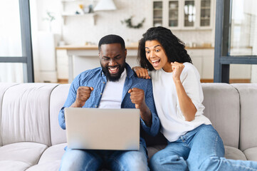 Hilarious African-American couple celebrates success raised clenched fists and screaming happily, sits on the couch at home with the laptop on the laps, won in lottery, got best offer