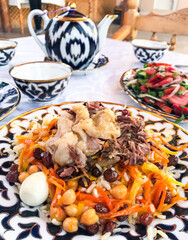 A set table in an Uzbek restaurant: wedding pilaf made of rice with carrots. With lamb and tail fat. As well raisins, chickpeas and eggs. On blurry background, a tomato salad and a teapot with bowls