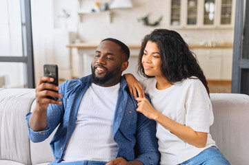 Obraz premium African-American couple with smartphone on the couch spends leisure time in social networks, scrolling feed, watching videos, a black guy is showing smth to his girlfriend