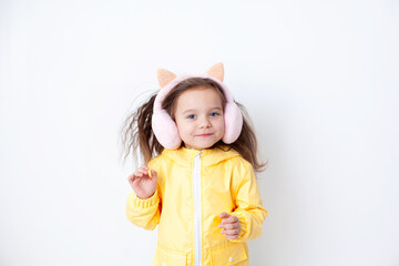 A cute girl in clothes for spring or autumn jumps and smiles. Yellow jacket and fluffy headphones. White background, place for text.
