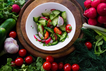 A pile of fresh, ripe vegetables: radishes, tomatoes, cucumbers, herbs, and garlic are arranged around a round wooden chopping board. On it is a white bowl with vegetables cut into a salad