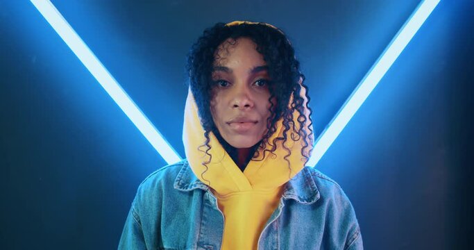 Handheld portrait of stylish african american black woman in yellow hoodie and jeans jacket standing in night disco club and looking at camera backlit with blue neon light stripes. Gen z urban female