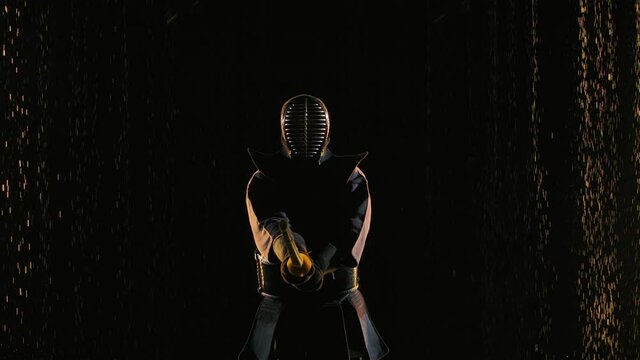 Japanese kendo fighter in fighting stance holding your shinai. Athlete in traditional martial armor and with bamboo sword in hands poses against black studio background and rain. Close up. Slow motion