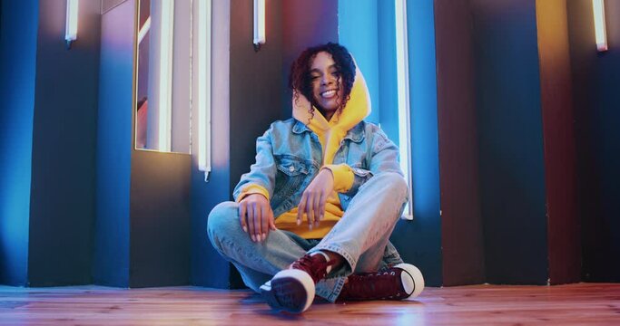 Stylish cheerful african american woman in yellow hoodie and jeans jacket listening music and dancing sitting by colorful neon light stripes background in night disco club. Gen z urban female