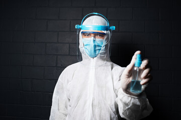 Portrait of doctor wearing PPE suit against coronavirus and covid-19, holding sanitizer bottle spray on background of black brick wall.