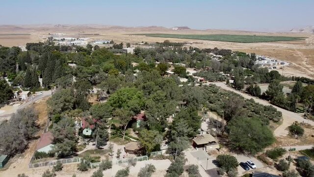 Aerial backwards panoramic drone shot of kibbutz Sde-Boker, filled with trees, bushes, fields, trails, buildings and houses.  A view of mountains and blue sky in the horizon. Day.