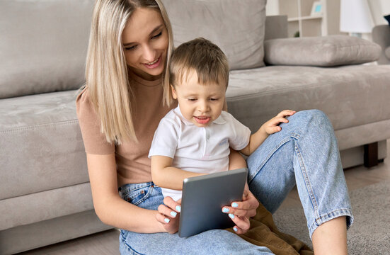 Young mom sitting at home on floor with kid of 3-4 years old, watching online activities classes for early education on tablet computer, showing movies for kids.
