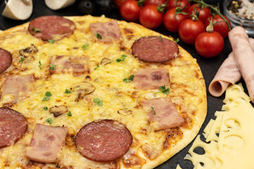 Pizza with ham, salami and mushrooms on a black background close-up with ingredients.