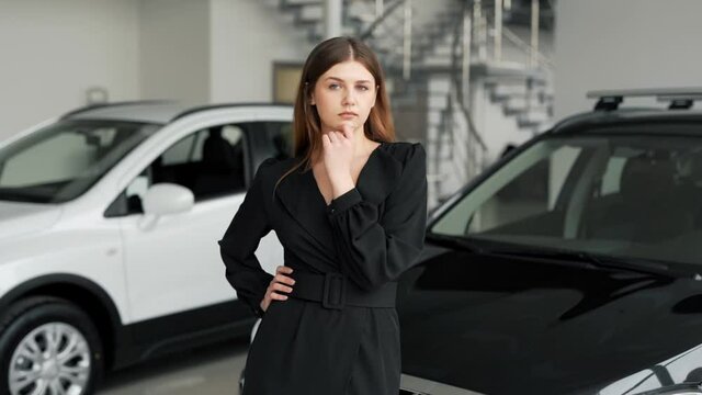 Beautiful girl in a black dress stands on a background of cars pondering over buying a car