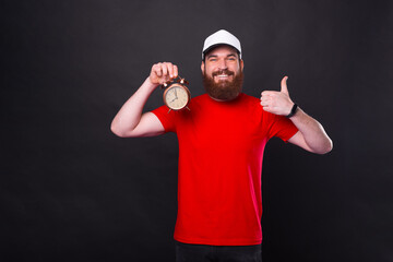 Good time manager over black background is holding an alarm clock and showing like gesture.