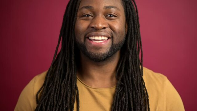 Friendly and positive African-American with dreadlocks guy wearing bright t-shirt looks at camera and smiles isolated on red background, happy dark skinned man feels good, has lucky day today