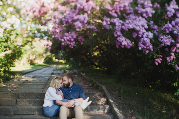 Romantic and happy caucasian couple in casual clothes hugging on the background of beautiful blooming lilac. Love, relationships, romance, happiness concept. Man and woman walking outdoors together.