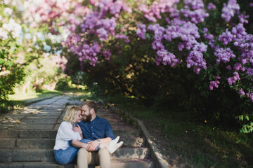 Fototapeta premium Romantic and happy caucasian couple in casual clothes hugging on the background of beautiful blooming lilac. Love, relationships, romance, happiness concept. Man and woman walking outdoors together.