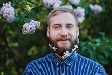 Young smiling caucasian man with lilac flowers in his beard having fun and posing for a photo.