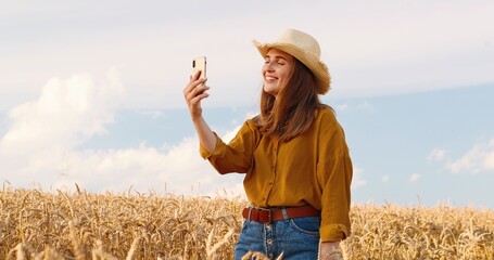 Beautiful Caucasian happy girl in hat having video conference on smartphone while standing in wheat field in village. Pretty young female videochatting on cellphone outdoors. Video call concept