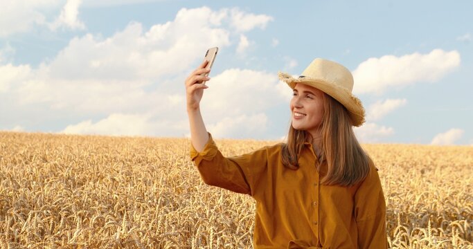 Portrait of beautiful happy Caucasian girl making selfie photo on smartphone while standing in golden field. Pretty joyful young woman in hat taking pictures in countryside. Village concept