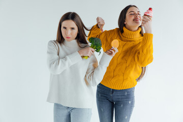 Hard choice. Two young sporty girls is choosing between fruits and sweets while standing on gray background. Junk food or healthy food?