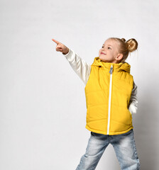 pretty smiling blonde girl in a yellow puffy vest is standing with her hand in her pocket and pointing with her other hand. Side view
