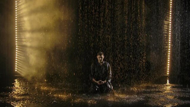 Portrait of a man in a black kimono sitting on the surface of the water and meditating in the rain. Japanese kendo fighter posing against dark smoky studio background with yellow light. Slow motion.