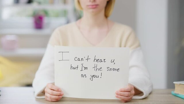 Close-up of I can't hear u but I'm the same as you written on paper with blurred smiling unrecognizable young Caucasian woman at background. Deaf disabled positive lady posing indoors