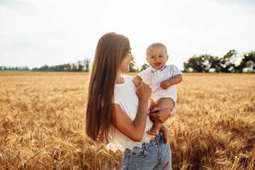 A mother holds her son on the hands at the golden wheat field. Woman with her child spend witme together outside having fun and enjoying good day. Family values and joy concept,