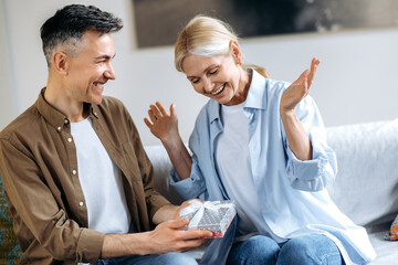 Happy middle aged couple in love. A loving joyful gray-haired husband gives a beautiful box with an expensive gift for their marriage anniversary or her birthday, charming wife is happy and smiling