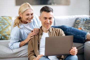 Happy caucasian family couple, mature wife and husband at home on a sofa, in casual wear, using laptop, browsing internet, social networks, chatting with friends or family, looking for household goods