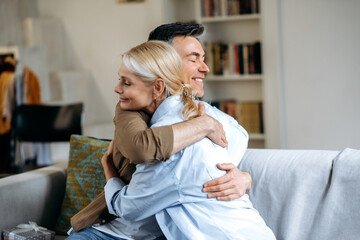 Happy loving senior caucasian family, happy husband and wife dressed in casual stylish clothes, sitting at home on sofa in living room, hugging, eyes closed, spend time together, smiling