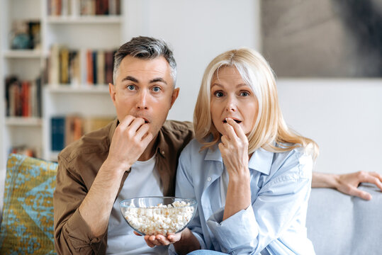 Middle aged shocked caucasian family couple sitting at home on sofa in casual clothes, watching tv show or exciting movie, eating popcorn, spending weekend together at home