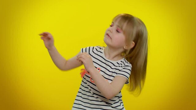 Trendy cheerful positive little blonde teen kid child girl having fun dancing and moving to rhythm, dabbing raising hands, making dubdance gesture. Yellow studio background. Young children emotions