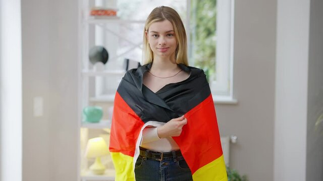Portrait of confident young woman wrapping in German flag looking at camera smiling. Happy relaxed Caucasian lady posing with national symbol at home indoors. Pride and patriotism concept