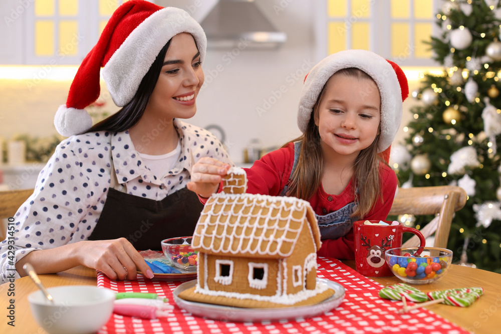 Sticker Mother and daughter decorating gingerbread house at table indoors