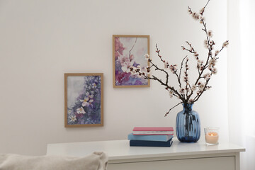 Flowering tree twigs in glass vase, burning candle and books on table near white wall at home