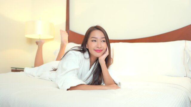 Happy pretty Asian woman lying on her belly on the hotel made-up bed wearing a white dressing gown, smiling front view