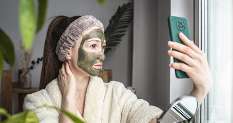 A young pretty woman in a bathrobe and with a green cosmetic mask on her face is taking a selfie on her mobile phone