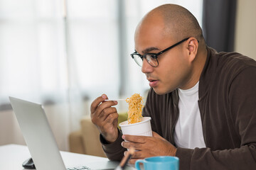 Young asian businessman working with laptop. Indian freelancer eating instant noodle while working. He work at home. He had to hurry and finish his work.