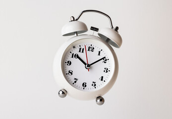Small white alarm clock, black numbers, set the time for 10.10 o'clock, placed on a white table.