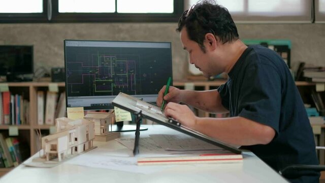 An Asian architect  in black T-shirt  is working on a design in his studio using a computer-aided design program and drafting board to drafting the construction plan.