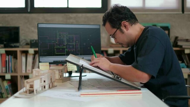 An Asian architect in a black T-shirt is working on a design work in his studio using a computer-aided design program and drafting board to drafting the construction plan.