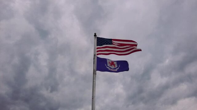 Flags of the United States of America and Talbot County, Maryland are flying together on a flag post against cloudy sky. Slow motion footage.