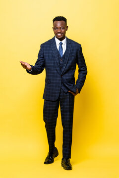 Full lenght portrait of young handsome African man in business suit smiling and opening his palm in yellow isolated studio background