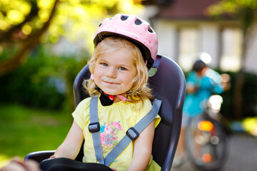 Fototapeta na wymiar Portrait of little toddler girl with security helmet on the head sitting in bike seat of parents. Boy on bicycle on background. Safe and child protection concept. Family and weekend activity trip.