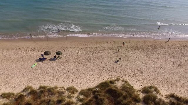 Steady aerial shot of a coastline . Sea shore and vegetation. Some people on the beach with a couple of umbrellas and fishermen with some lines set.