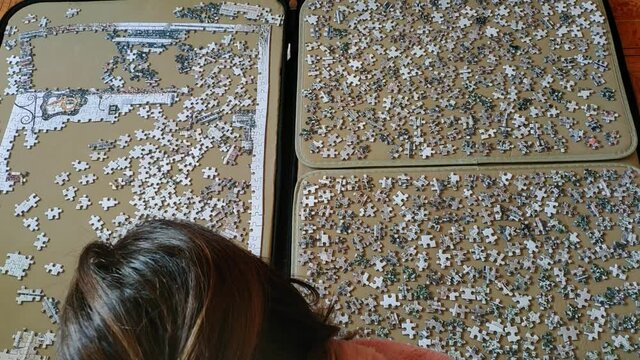 Top view of a woman on comfortable clothes at hme working on a big puzzle. The right hand side is full of the spare pieces and on the left hans side is the beginning of the puzzle being made.
