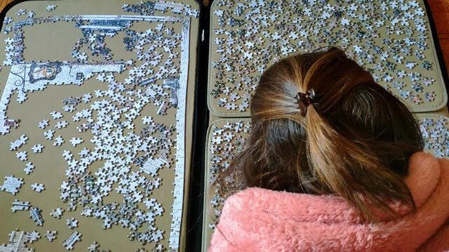 Top view of a woman on comfortable clothes at hme working on a big puzzle. The right hand side is full of the spare pieces and on the left hans side is the beginning of the puzzle being made.