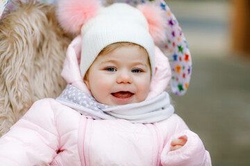 Fototapeta na wymiar Cute little beautiful baby girl sitting in the pram or stroller on cold autumn, winter or spring day. Happy smiling child in warm clothes, fashion stylish baby coat and hat. Snow falling down