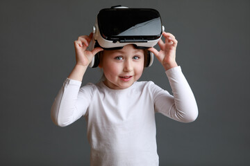 Smiling little kid girl in white long sleeve shirt isolated on grey background wearing VR glasses