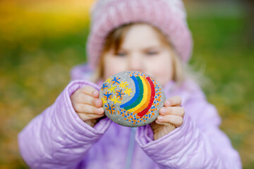 Adorable little toddler girl with painted rainbow on stone during pandemic coronavirus quarantine, outdoors. Child painting rainbows around the world as hope, sign and fight against covid corona virus